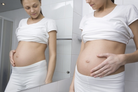 Pregnant woman stands looking at stomach in bathroom mirrorの写真素材
