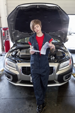 Young female mechanic reading paper with open bonnet of car in garageの写真素材