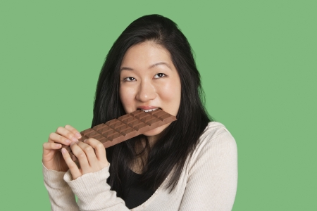 Cute young woman eating a large chocolate bar over green backgroundの写真素材