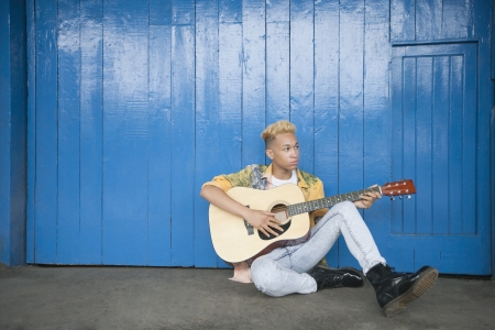 Trendy teenage boy playing guitar as he sits against wood paneled wallの写真素材