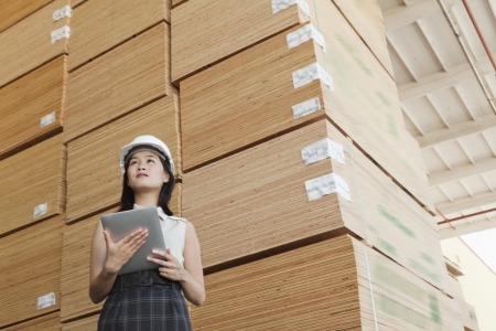 Low angle view of female industrial worker holding tablet PC with stacked wooden planks in backgroundの写真素材