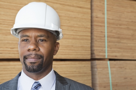 Confident African American male contractor looking away with stacked wooden planks in backgroundの写真素材