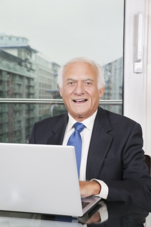 Portrait of happy elderly businessman with laptop sitting at tableの写真素材