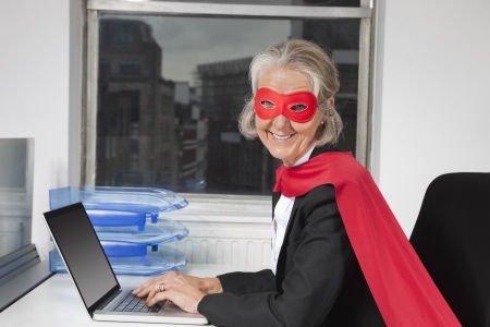 Portrait of senior businesswoman in superhero costume using laptop at office deskの写真素材