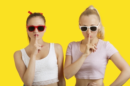 Portrait of two playful young women with fingers on lips over yellow backgroundの写真素材