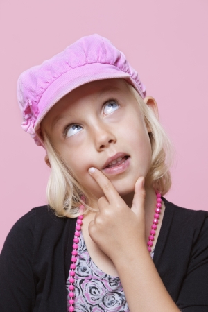 Thoughtful young girl wearing cap with finger on chin over pink backgroundの写真素材