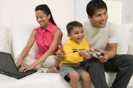 Father playing video game with son by mother using laptop on couch