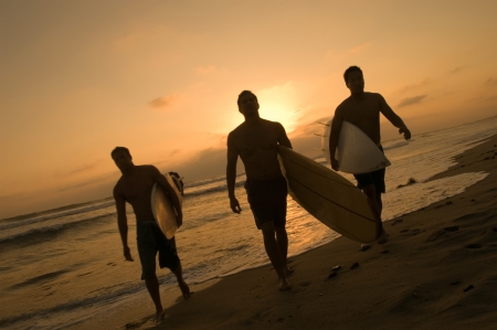 Three surfers carrying surfboards out of surf at sunsetの写真素材