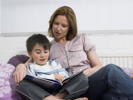 Mother and son (5-6) reading book on couch in living roomの写真素材