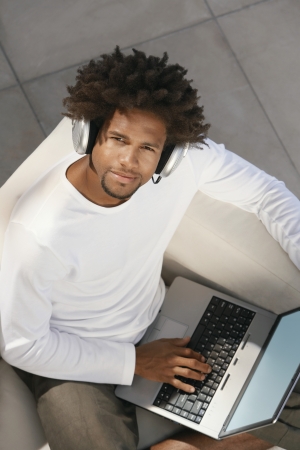 Young man sitting on sofa outside using laptop and listening to music through earphones portrait overhead viewの写真素材