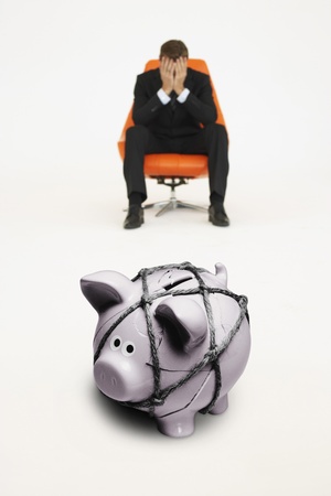 Worried businessman on chair and piggybank tied with rope representing financial difficultiesの写真素材