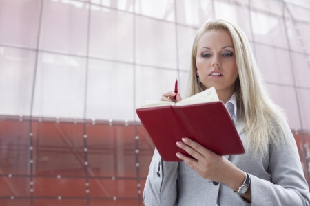 Beautiful businesswoman reading notes in organizer while standing against office buildingの写真素材