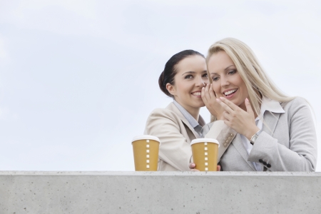 Happy businesswomen with disposable coffee cups sharing secrets against clear skyの写真素材