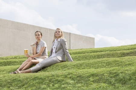 Full length of relaxed businesswomen in formals sitting on grass steps against skyの写真素材