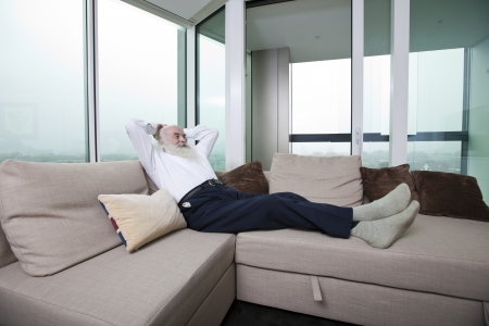Full length of senior man relaxing on sofa with hands behind head at homeの写真素材