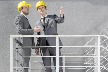 Two young businessmen in hard hats discussing on stairwayの写真素材