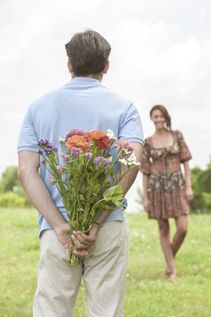 Rear view man surprising woman with bouquet in parkの写真素材