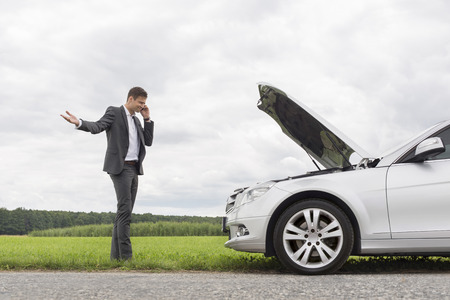 Frustrated young businessman using cell phone by broken-down car at countrysideの写真素材