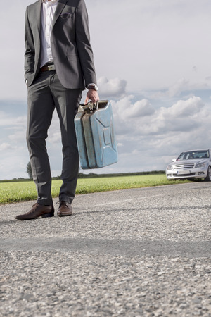Low section of young businessman carrying petrol can with broken car in background at countrysideの写真素材