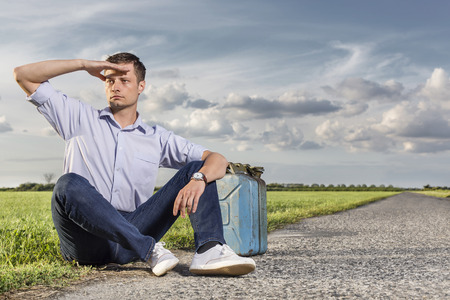 Full length of young man shielding eyes while sitting with petrol can by country roadの写真素材