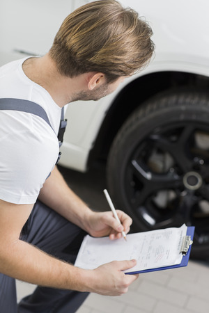 Side view of automobile mechanic writing on clipboard while examining car's wheel in workshopの写真素材