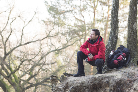 Full length of male hiker crouching on cliff in forestの写真素材