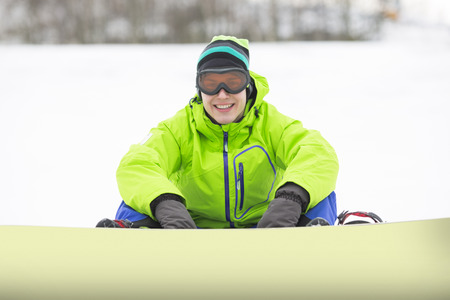Portrait of smiling young man with snowboard sitting on snowy landの写真素材