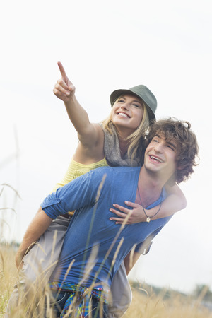 Cheerful woman showing something while enjoying piggyback ride on man in fieldの写真素材