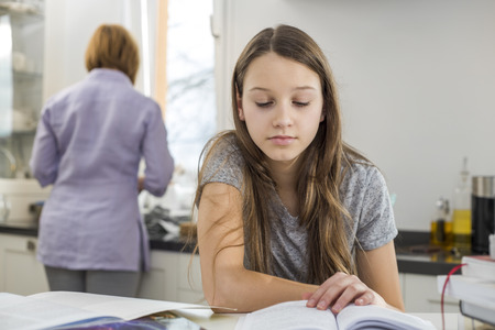 Girl studying at table with mother standing in backgroundの写真素材