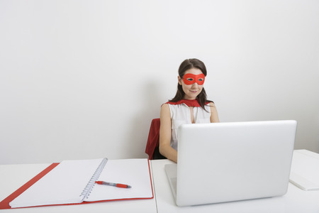 Young businesswoman dressed as superhero using laptop at desk in officeの写真素材