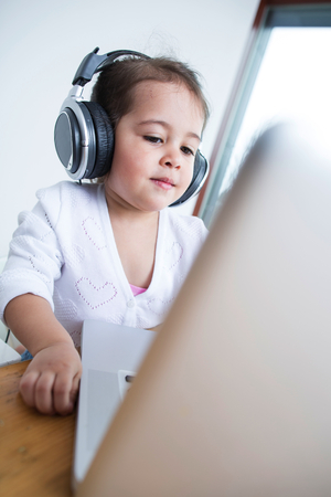 Little girl wearing headphones while looking at laptop at table in houseの写真素材