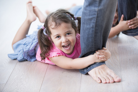 Portrait of playful girl with sister holding father's legs on hardwood floorの写真素材
