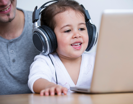 Little girl wearing headphones while looking at laptop with father at table in house