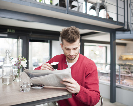 Young man reading newspaper while drinking coffee in cafeの写真素材