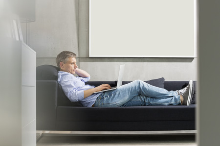 Full-length side view of Middle-aged man using laptop while lying on sofaの写真素材