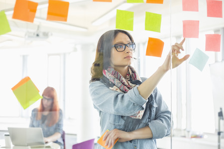 Creative businesswoman reading sticky notes on glass wall with colleague working in background at officeの写真素材