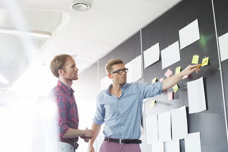 Creative businessmen discussing over sticky paper on wall in office