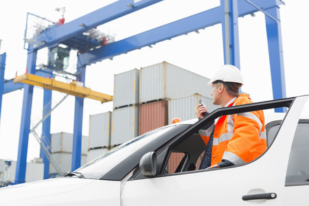 Middle-aged man using walkie-talkie while standing at car door in shipping yard