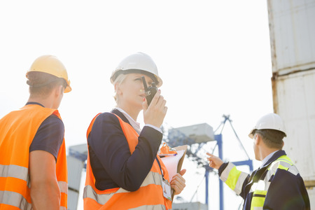 Female supervisor using walkie-talkie while workers discussing in background at shipyardの写真素材