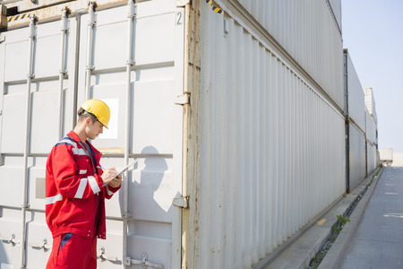 Male worker inspecting cargo container while writing on clipboard in shipping yard