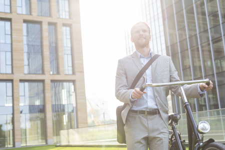 Thoughtful businessman standing with bicycle outside buildingの写真素材