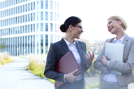 Cheerful businesswomen conversing while holding folder and laptop outside office buildingの写真素材