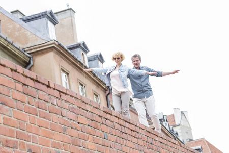 Low angle view of middle-aged couple with arms outstretched walking on brick wall against clear skyの写真素材