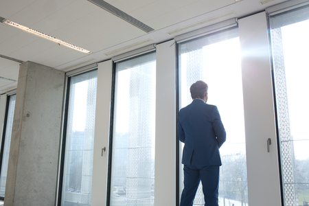 Rear view of mature businessman looking through office windowの写真素材