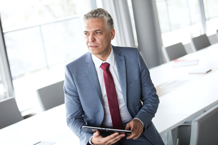 Thoughtful mature businessman holding digital tablet while leaning on conference tableの写真素材