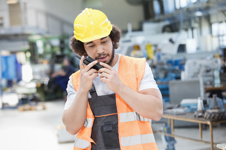 Young manual worker wearing protective clothing while using walkie-talkie in metal industry