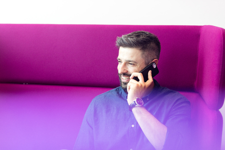 Smiling mid adult businessman talking on mobile phone while sitting on purple couch at officeの写真素材