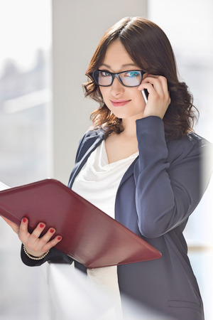 Portrait of smiling businesswoman using cell phone while holding file in officeの写真素材