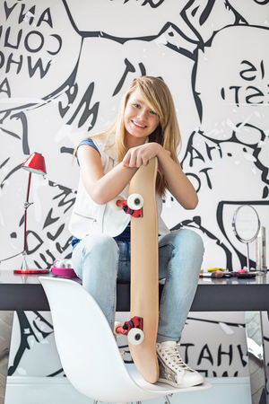 Full-length portrait of happy teenage girl with skateboard sitting on study table at homeの写真素材