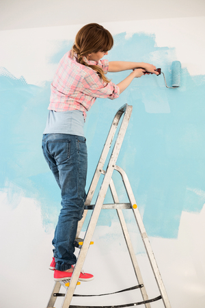 Woman on ladder painting wall with paint rollerの写真素材